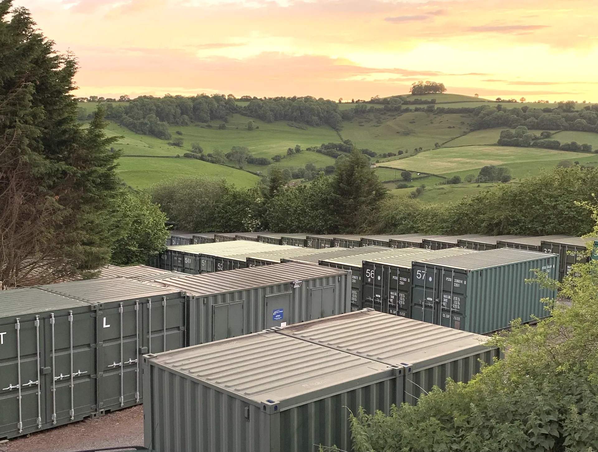 Rows of steel storage containers at Perry Storage's yard, with the Somerset countryside and a sunset behind